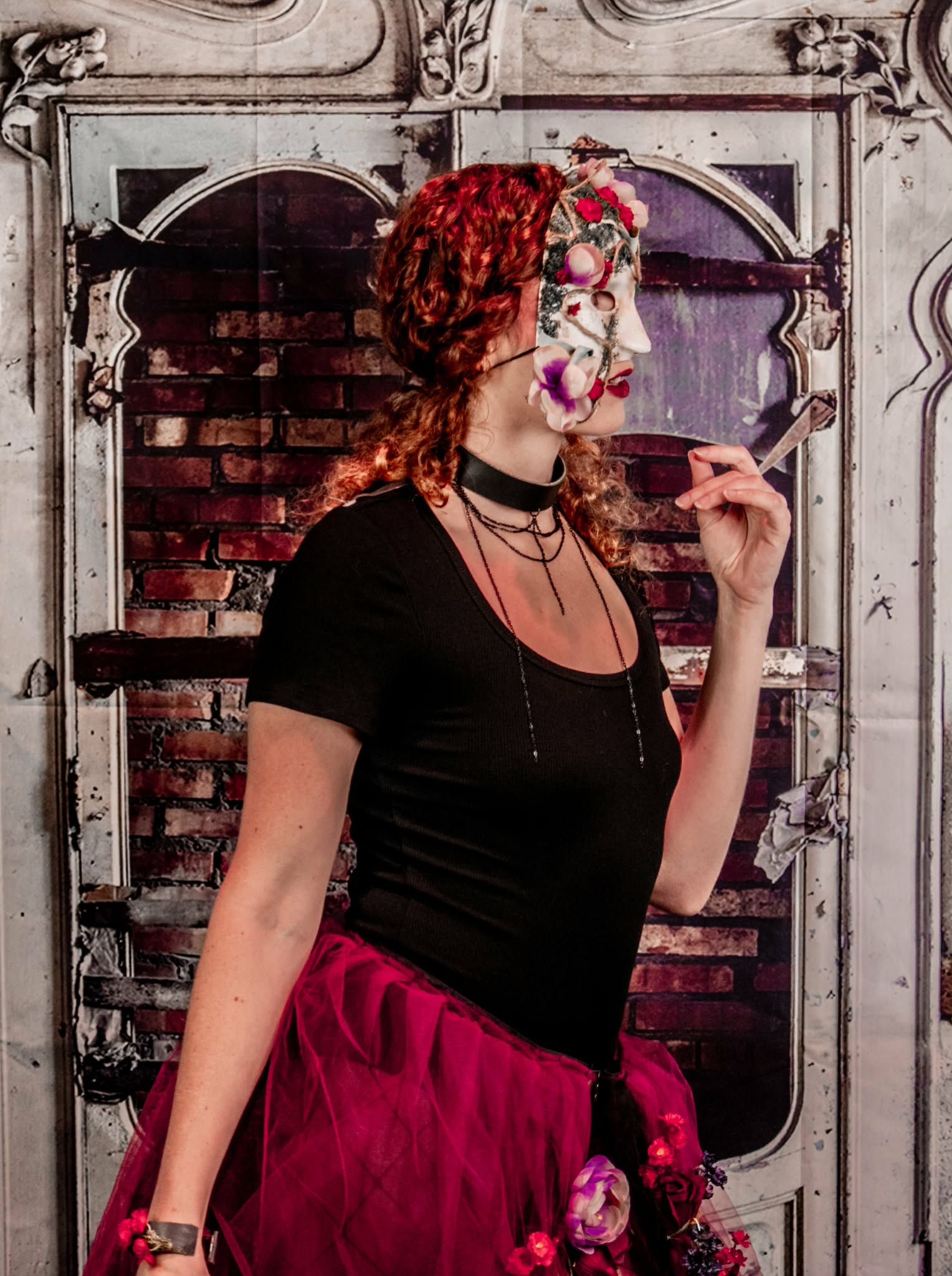 Profile wearing a distressed floral half-mask, layered chains, and red flower-covered skirt